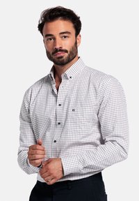 Man met een baard in een wit geruit overhemd met knopen die zijn manchetten aanpast, staande tegen een effen witte achtergrond.