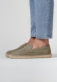 Mocasines de ante verde con suela de yute trenzado, que presentan un destacado detalle de costura en la parte superior y un diseño de punta redondeada y plana.