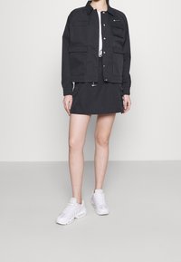 Czarna kurtka z wieloma kieszeniami, w parze z dopasowaną spódnicą i białymi trampkami. Strój cechuje się czystym, minimalistycznym designem i fakturą.