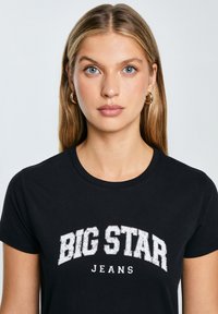 Juodas medvilninis marškinėlis trumpomis rankovėmis, su baltu grafiniu dizainu, kuriame užrašyta "BIG STAR JEANS" nusidėvėjusiomis raidėmis per krūtinę.