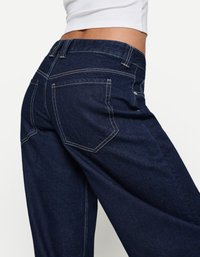 Jean en denim bleu foncé avec poches arrière, coutures blanches contrastantes et coupe droite. La texture semble lisse avec un léger éclat.
