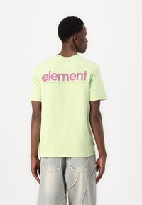 Světle zelené bavlněné tričko s velkým růžovým logem "element" na zadní straně. Model má na sobě vybledlé šedé volné džíny.