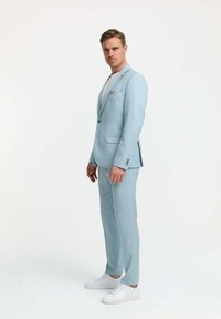 Hellblauer Anzug mit einem maßgeschneiderten Blazer und passenden Hosen, ergänzt durch ein weißes T-Shirt und weiße Sneaker. Bemerkenswertes Taschentuch-Detail.