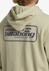 Sudadera beige con un gran logotipo gráfico en azul oscuro con acentos en rojo y azul, que muestra el texto "billabong" y "boardshorts".