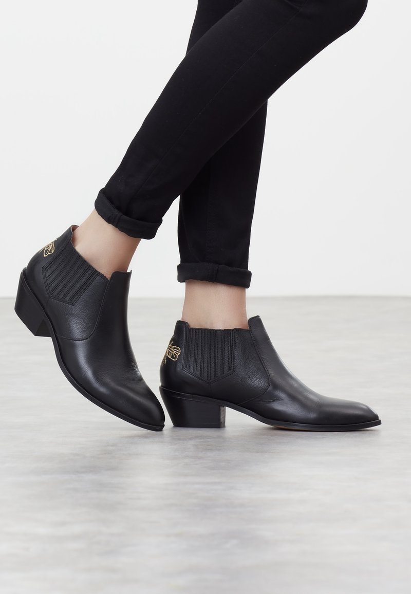 Personne portant des bottines en cuir noir à petits talons carrés et une broderie dorée d'abeille sur les côtés, associées à un pantalon noir retroussé.