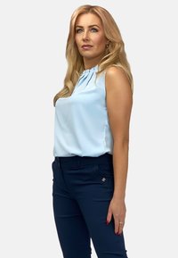 Jasnoniebieska bezrękawnikowa bluzka z pliskami przy dekolcie, zestawiona z ciemnonawojskowymi spodniami o dopasowanym kroju z bocznymi kieszeniami i dekoracyjnym akcentem.