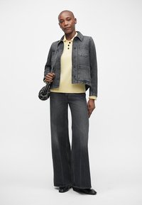 Woman wearing a black denim jacket, yellow buttoned sweater, wide-leg black jeans, and holding a black studded handbag against a plain background.
