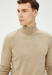 Beige Rollkragensweater aus weichem Strickmaterial, mit tailliertem Schnitt und gerippten Ärmelbündchen sowie Kragen, vor einem schlichten Hintergrund.