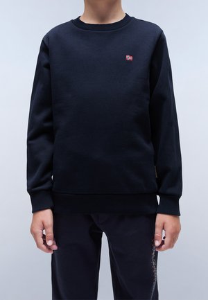 Barn iført en marineblå sweatshirt med et lille norsk flagemblem på øvre venstre bryst, kombineret med mørke bukser.