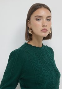 Junge Frau mit glattem braunem Haar, die einen strukturierten dunkelgrünen Pullover und große, goldene, gedrehte Creolen trägt, vor einem schlichten Hintergrund.