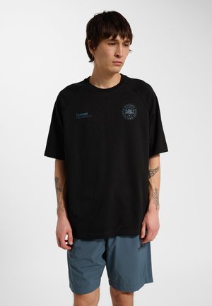 Mand med tatoveringer iført sort sportst-shirt og blå shorts, står med afslappede hænder mod en ensfarvet lys baggrund.