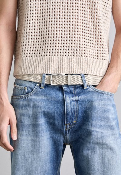 Beige gebreid mouwloos topje met open geweven textuur, gecombineerd met lichtblauwe denim jeans en een gevlochten lichtgrijze riem met een zilveren sluiting.