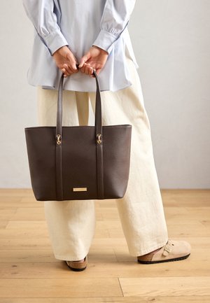 Personne portant un pantalon beige clair et des chaussures à enfiler beige tenant un sac fourre-tout en cuir brun foncé derrière son dos.