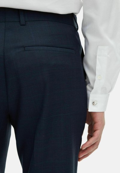 Granatowe, dopasowane spodnie w delikatny wzór w kratkę i boczną kieszenią. Częściowo widoczna biała koszula z mankietami zapinanymi na guziki.