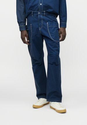 Blå denimbukser med en afslappet pasform, der har hvid søm, flere lommer og et flagrende bendesign. Kombinationen med hvide sneakers.