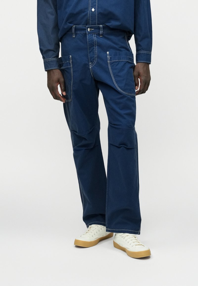 Μπλε τζιν παντελόνι με άνετη εφαρμογή, με λευκές ραφές, πολλές τσέπες και σχέδιο καμπάνα στα πόδια. Συνδυασμένο με λευκά αθλητικά παπούτσια.