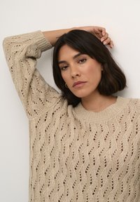 Maglione beige a maglia con un motivo in pizzo ondulato, maniche corte e colletto a coste. La texture è morbida e traspirante.