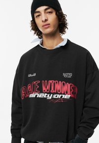 Černá mikina s oversized střihem, s grafickým textem "VÍTĚZ ZÁVODU" v červené a bílé barvě a vyšívanými detaily loga na hrudi a rukávu.