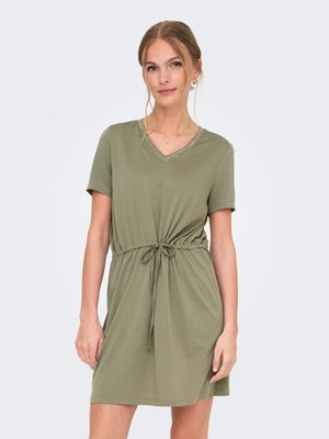 Jeune femme portant une robe olive à manches courtes avec un col en V et une taille ajustable par un cordon, debout devant un fond clair uni.