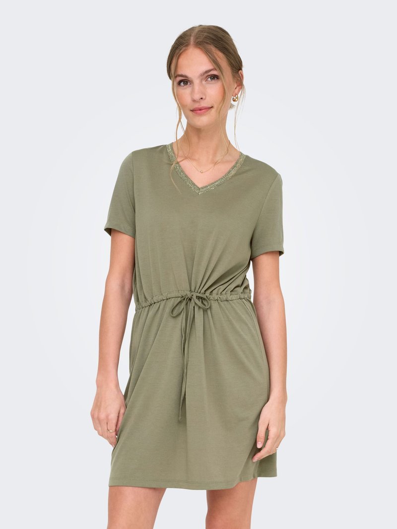 Jeune femme portant une robe olive à manches courtes avec un col en V et une taille ajustable par un cordon, debout devant un fond clair uni.