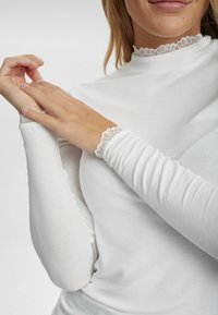 Witte geribbelde lange mouwen top met een hoge halslijn en kantafwerking langs de kraag en manchetten. Zacht materiaal met een aansluitend ontwerp.