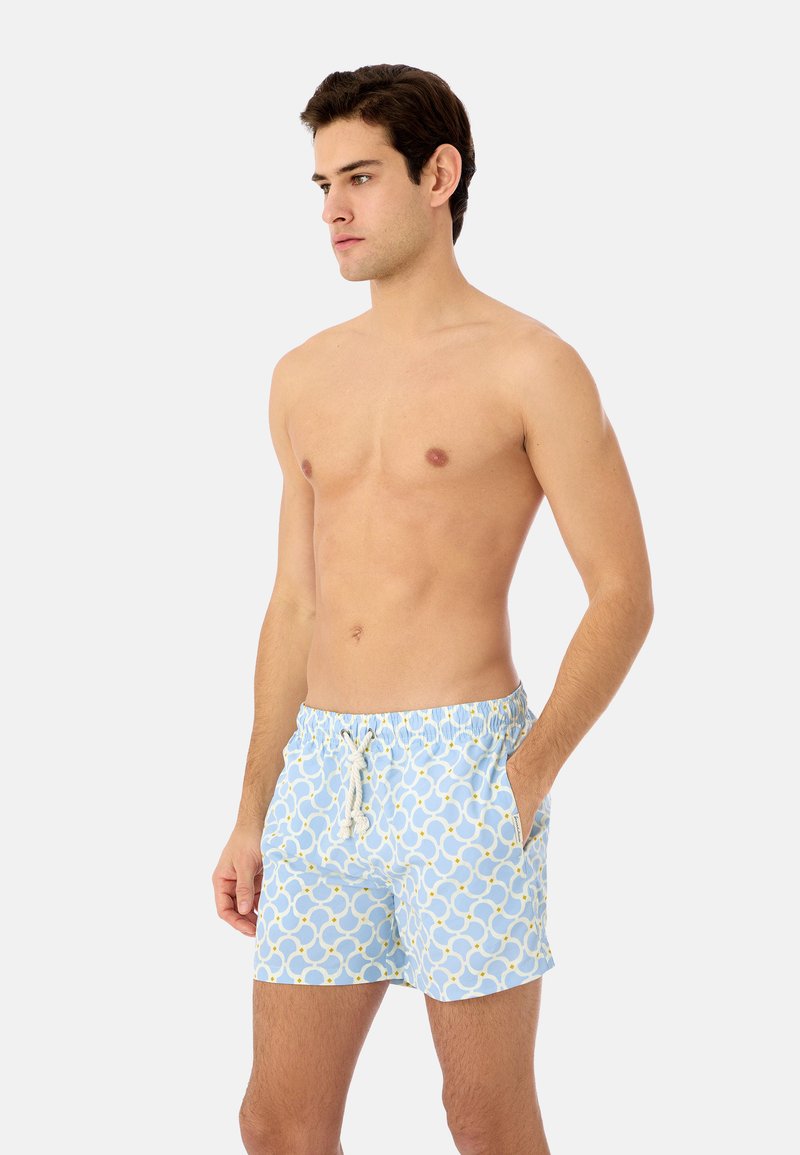 Jeune homme aux cheveux courts et foncés portant un short de bain bleu clair à motif avec un cordon blanc, debout avec une main dans la poche.