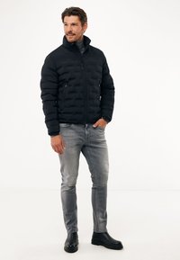 Černá péřová bunda s texturovaným designem, vysokým límcem, zipovými kapsami, nošená přes šedý rolák, v kombinaci se šedými džíny a černými botami.