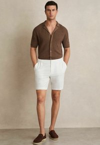 Cămașă scurtă maro cu model hexagonal, pantaloni scurți albi cu talie elastică și pantofi slip-on maro. Outfit simplu de vară.