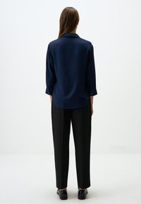 Blusa di seta blu navy con colletto e maniche tre quarti, abbinata a pantaloni neri larghi e scarpe nere, vista da dietro.