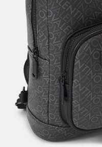 Gray backpack featuring a textured surface with a repeating logo pattern, black accents, and multiple zippered compartments for storage.