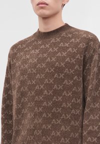 Homme portant un pull en tricot marron avec un motif répétitif "AX" marron clair et un col rond côtelé.