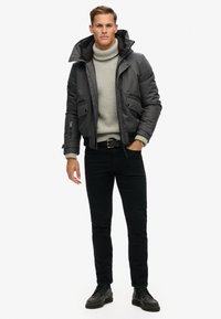 Grijze jas met een hoge kraag, voorzakken en geribbelde manchetten, gedragen over een lichtgrijze coltrui, gecombineerd met zwarte broek en schoenen.