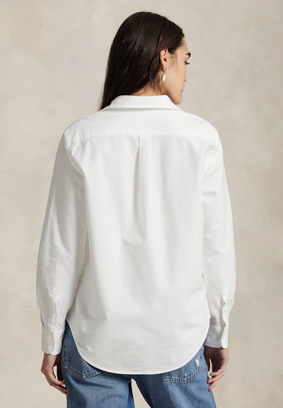 Kobieta z długimi ciemnymi włosami nosi białą koszulę z guzikiem i niebieskie dżinsy, pokazana od tyłu na neutralnym tle.