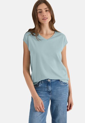Jeune femme aux cheveux châtain clair portant un haut bleu clair à col en V et manches courtes, ainsi qu'un jean bleu, posant devant un fond uni.