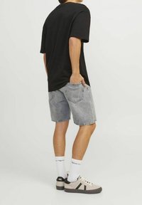 Persona vestida con una camiseta negra, pantalones cortos de mezclilla claros, calcetines blancos con texto y zapatillas beige con rayas negras, de pie contra un fondo blanco.