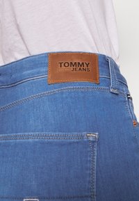 Jeans de mezclilla azul con una etiqueta de cuero marrón en la cintura que presenta el texto en relieve "TOMMY JEANS", con costuras visibles y un bolsillo trasero.