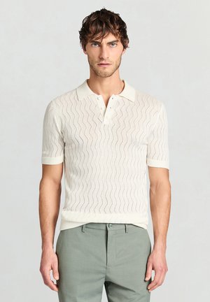 Hellcremefarbenes Polo mit kurzen Ärmeln und Wellenmuster, ausgestattet mit einer Dreiknopfblende und gerippten Bündchen, kombiniert mit hellgrünen Hosen.