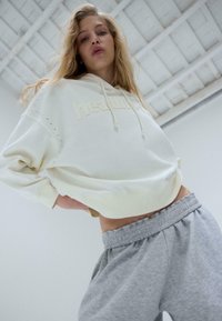 Sweat à capuche crème avec lettres en relief et accents en strass, associé à un pantalon de survêtement gris à taille élastique. Tissu lisse, coupe contemporaine.