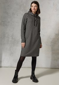 Schwarzes und weißes Hahnentritt-Musterkleid mit einem Cowl-Neck, langen Ärmeln und zwei seitlichen Taschen, kombiniert mit schwarzen klobigen Stiefeln.