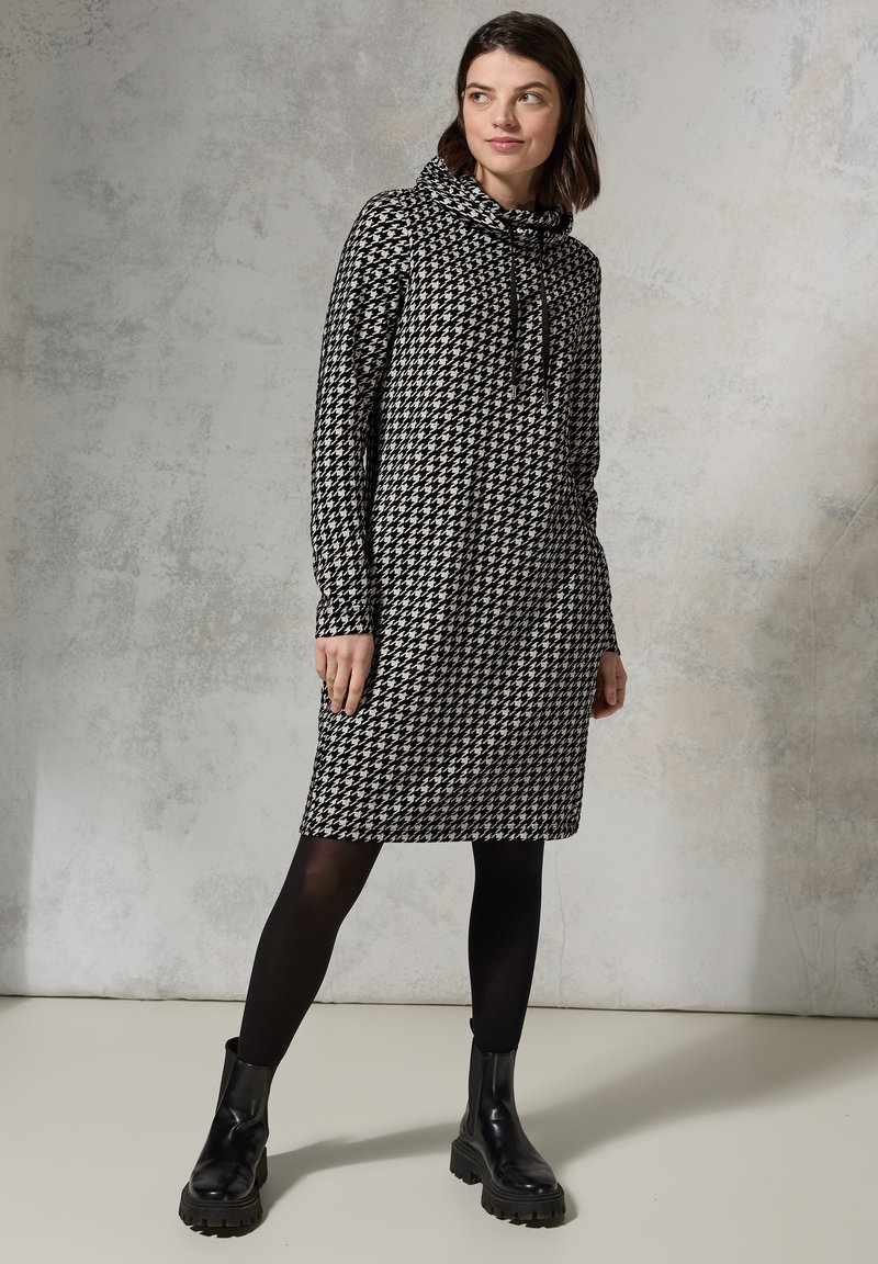 Schwarzes und weißes Hahnentritt-Musterkleid mit einem Cowl-Neck, langen Ärmeln und zwei seitlichen Taschen, kombiniert mit schwarzen klobigen Stiefeln.