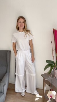 Λευκό μπλουζάκι και φαρδύ λευκό παντελόνι φόρμας με μαύρες πλευρικές ρίγες, απαλή υφή, άνετη εφαρμογή, σε συνδυασμό με λευκά αθλητικά παπούτσια. Το φόντο περιλαμβάνει φυτά.