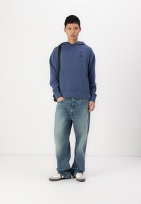 Μπλε φούτερ με μπροστινή τσέπη και λογότυπο, συνδυασμένο με χαλαρά ανοιχτόχρωμα τζιν και γκρι αθλητικά παπούτσια. Το μοντέλο στέκεται μπροστά σε λευκό φόντο.