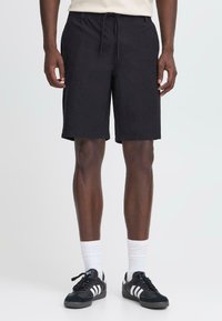Shorts en coton noir avec une taille élastique et un cordon de serrage. Coupe au genou ; associés à des baskets noires et des chaussettes blanches.