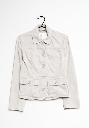 Veste beige clair à boutons avec col et deux poches rabats à l'avant, suspendue sur un cintre noir sur fond blanc.