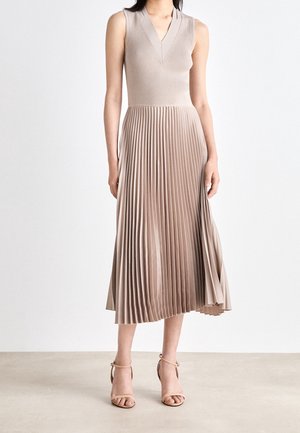 Kvinde iført ærmeløs beige V-hals top med plisseret midinederdel og nøgenfarvede ankelremssandaler, stående på neutral gulv mod hvid baggrund.