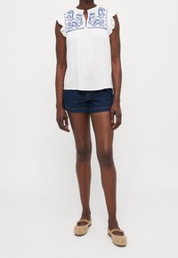 Blusa bianca con design ricamato blu sul colletto, maniche corte a volant, abbinata a pantaloni shorts in denim scuri e scarpe flat beige.