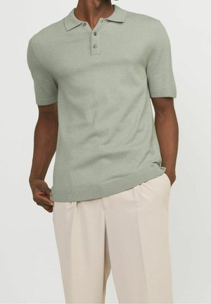 Homme portant un polo vert clair à manches courtes et un pantalon beige, une main tenant l'ourlet du polo et l'autre dans la poche.