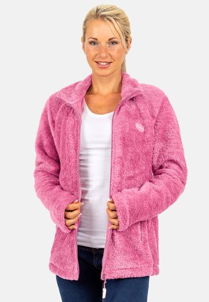 Vrouw draagt een roze zachte jas met rits over een wit shirt, houdt de randen van de jas met beide handen vast, glimlacht en kijkt recht vooruit.