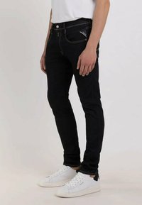 Jean slim noir avec surpiqures jaunes, design à cinq poches et zip, associés à des baskets blanches avec des accents noirs.