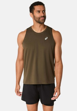 Homme athlétique portant un débardeur vert olive et un short noir, debout et regardant sur le côté devant un fond uni.